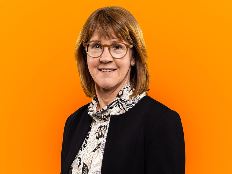 Porträt von Dr. oec. Constanze Fisch lächelnd vor einem leuchtend orangefarbenen Hintergrund.