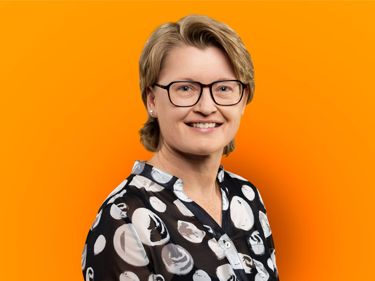 Porträt einer lächelnden Frau mit kurzen, blonden Haaren und Brille vor einem leuchtend orangefarbenen Hintergrund. Sie trägt eine schwarz-weiß gemusterte Bluse.