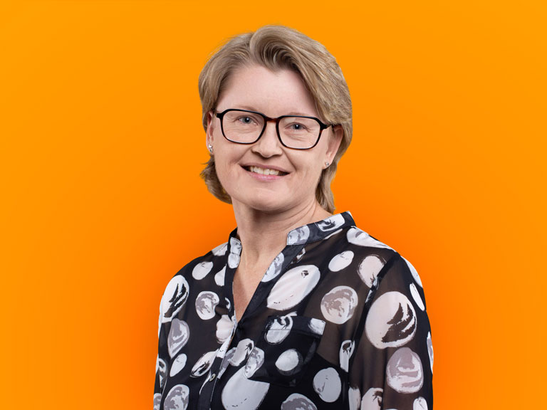 Porträt einer lächelnden Frau mit kurzen, blonden Haaren und Brille vor einem leuchtend orangefarbenen Hintergrund. Sie trägt eine schwarz-weiß gemusterte Bluse.