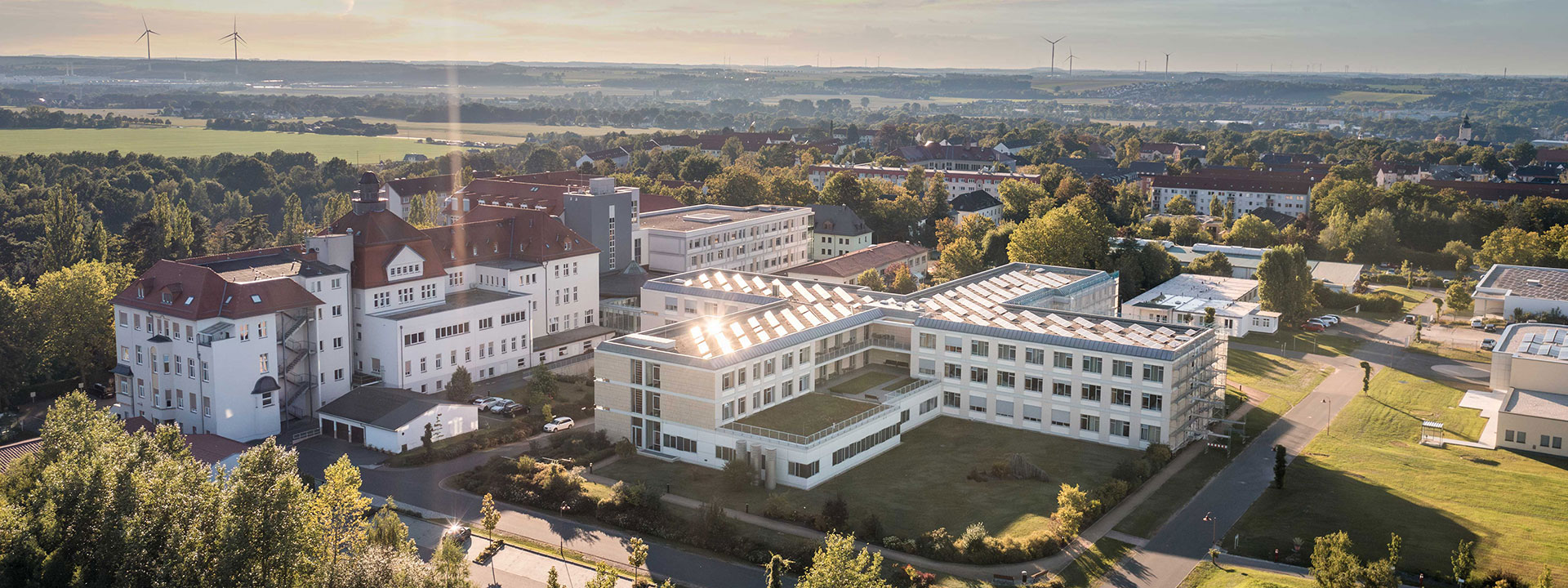 Luftaufnahme des Klinikum Glauchau bei Sonnenlicht, das den modernen Anbau und das historische Hauptgebäude zeigt. Im Hintergrund ist die umliegende Stadtlandschaft zu sehen.