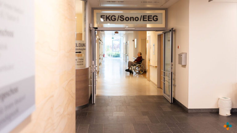 Eingang zu einer Klinikabteilung mit der Beschriftung „EKG/Sono/EEG“. Im hell beleuchteten Flur sitzt ein Patient im Rollstuhl.