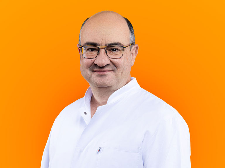 Porträt von Athanasios Karagiannis in weißer Berufsbekleidung lächelnd vor einem orangefarbenen Hintergrund.