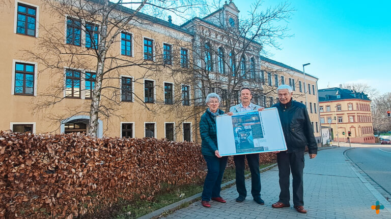 Mitglieder des Denkmalvereins Glauchau präsentieren eine Informationstafel vor einer historischen, gelb-beigen Schulanlage mit großen Fenstern. Im Vordergrund befindet sich eine herbstliche Hecke, im Hintergrund ist eine Straße mit Ampelkreuzung in Glauchau zu sehen.