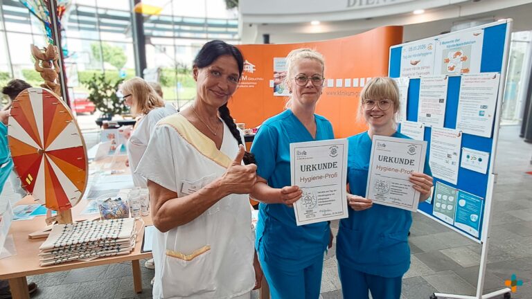 Drei Mitarbeiterinnen des Klinikums Glauchau präsentieren stolz ihre Urkunden als „Hygiene-Profi“ am Welthygienetag vor einem Infostand zur Händehygiene.