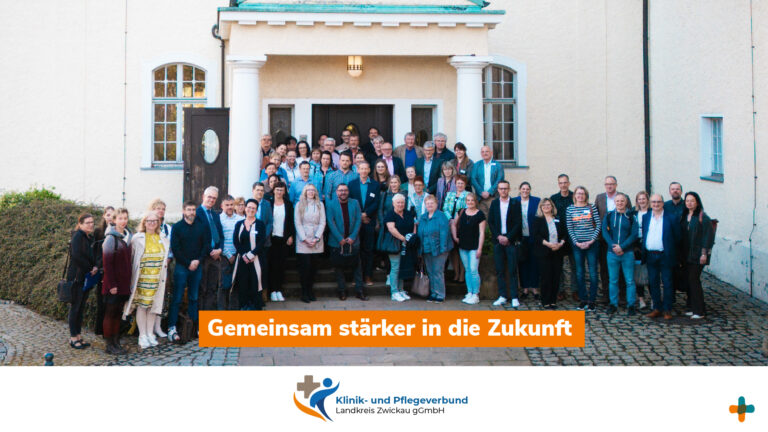 Anfrage erfolgreich Ein großer, lächelnder Gruppenbild-Verbund aus Männern und Frauen vor dem Eingang eines hellen Klinik- und Pflegegebäudes mit einem orangefarbenen Banner mit der Aufschrift „Gemeinsam stärker in die Zukunft“.