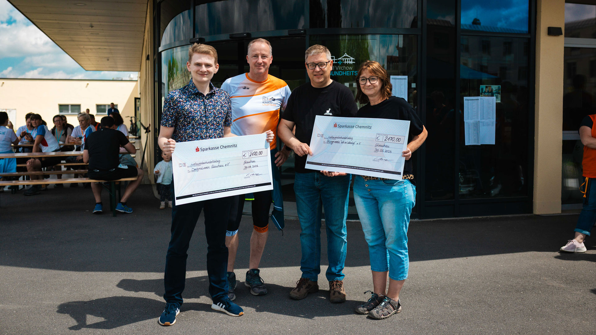 Gruppenfoto von einer Spendenübergabe: Zwei Männer und eine Frau halten zwei symbolische Schecks der Sparkasse Chemnitz vor einem Gesundheitszentrum. Links steht ein jüngerer Mann im gemusterten Hemd, daneben ein Mann in Sportkleidung. Im Hintergrund sind sitzende Menschen an Biertischen zu sehen.