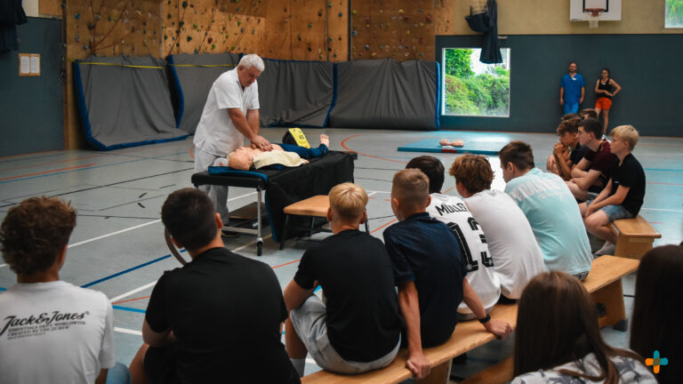 Ein Ausbilder demonstriert Jugendlichen in einer Sporthalle die Herzdruckmassage an einer Reanimationspuppe.