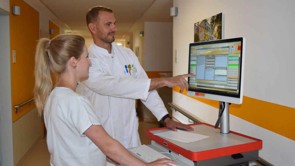 Ein Arzt und eine medizinische Fachangestellte besprechen Patientendaten an einem mobilen Computer-Bildschirm auf einem Klinikflur.