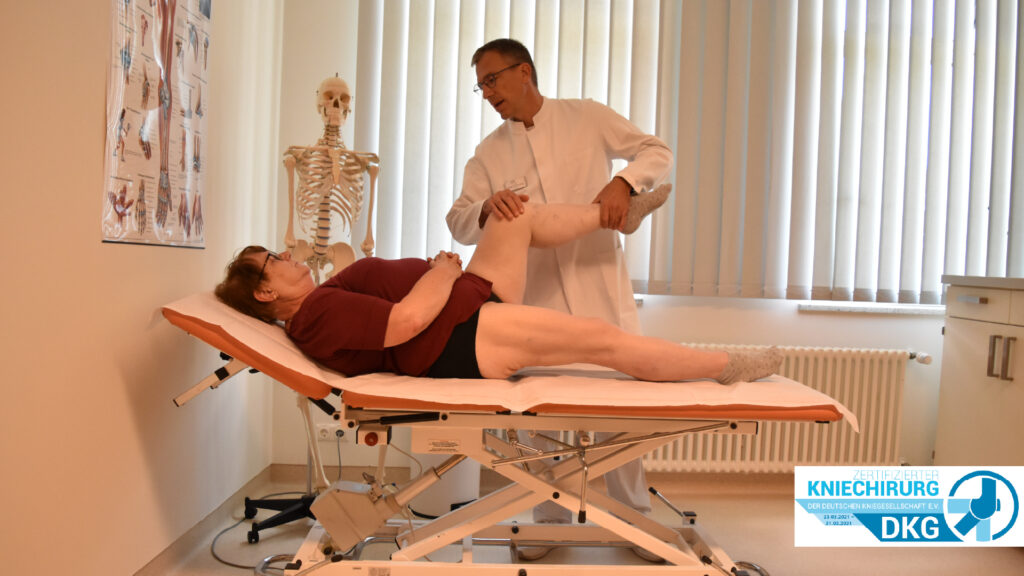 Ein Arzt untersucht das angewinkelte Knie einer auf einer Liege liegenden Patientin in einer orthopädischen Praxis.