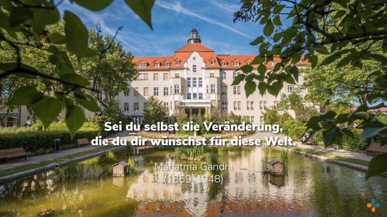 Großes, historisches Gebäude vom Klinikum Glauchau mit einem Teich im Vordergrund und dem Zitat von Mahatma Gandhi „Sei du selbst die Veränderung, die du dir wünschst für diese Welt.“