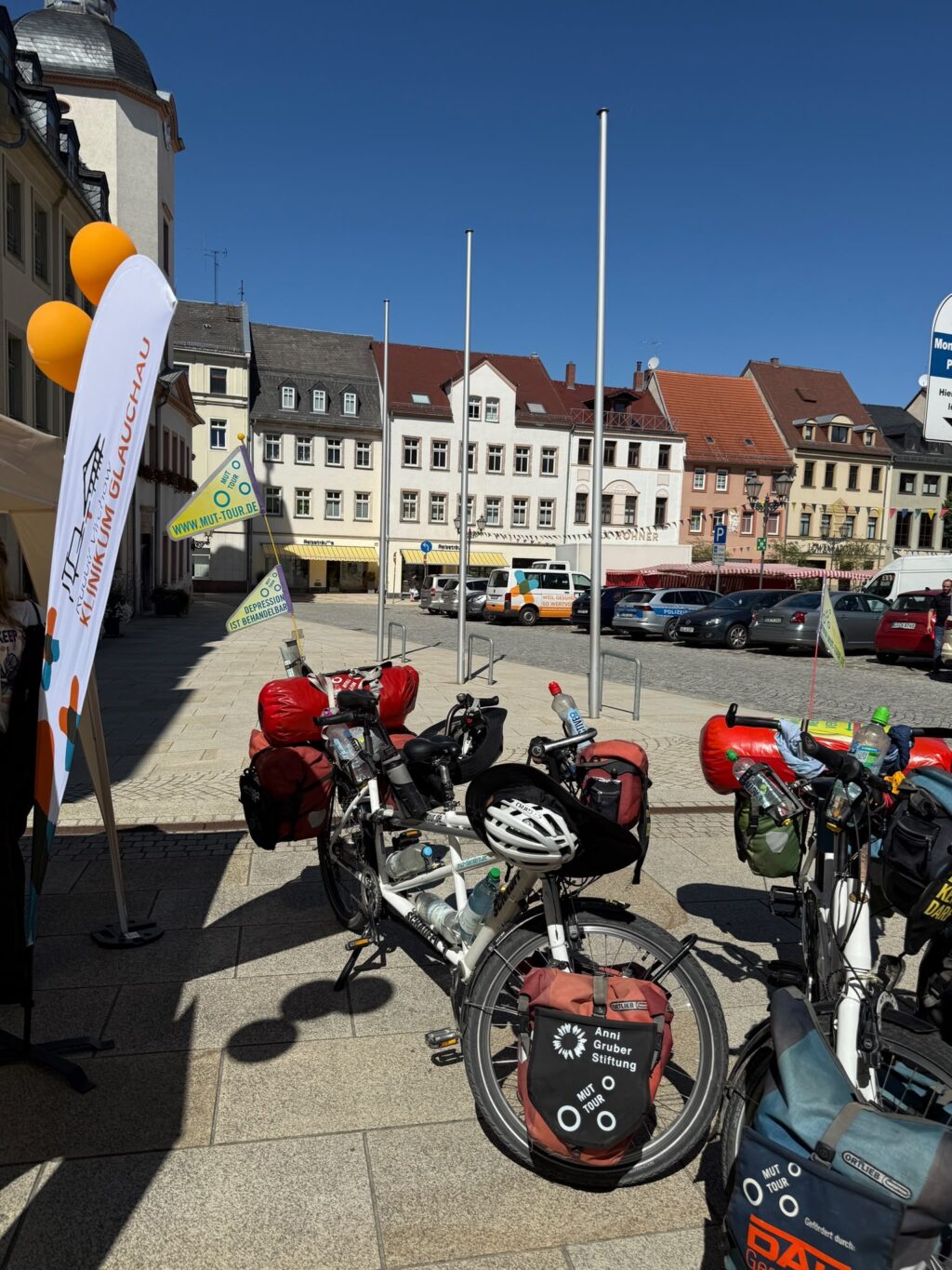 Bepackte Tandems der Mut-Tour auf einem sonnigen Marktplatz neben Werbefahnen des Klinikums Glauchau.