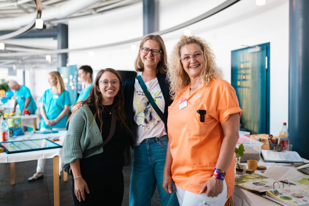 Drei lächelnde Frauen, darunter eine Mitarbeiterin vom Klinikum in orangener Arbeitskleidung, bei einer Klinikveranstaltung im Innenbereich.