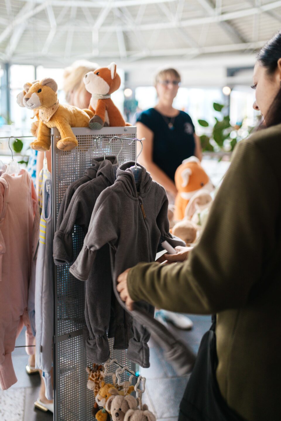 Frau betrachtet graue Baby-Overalls und Kuscheltiere an einem Verkaufsständer bei einer Baby-Messe.