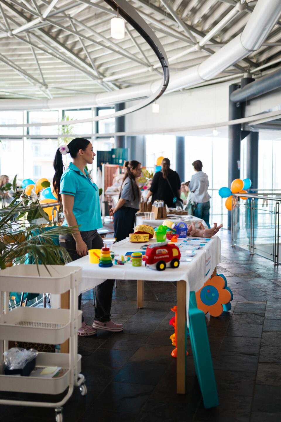 Mitarbeiterin an einem bunt dekorierten Informationsstand mit Spielzeug und Ballons in einem lichten Foyer.