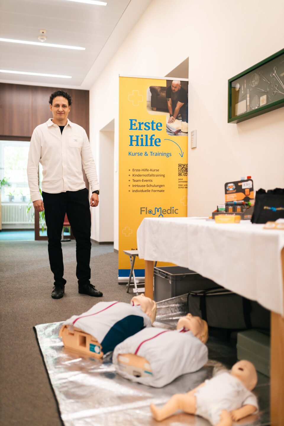 Mann steht neben einem Roll-up für Erste-Hilfe-Kurse mit Reanimationspuppen für Erwachsene und Babys am Boden.