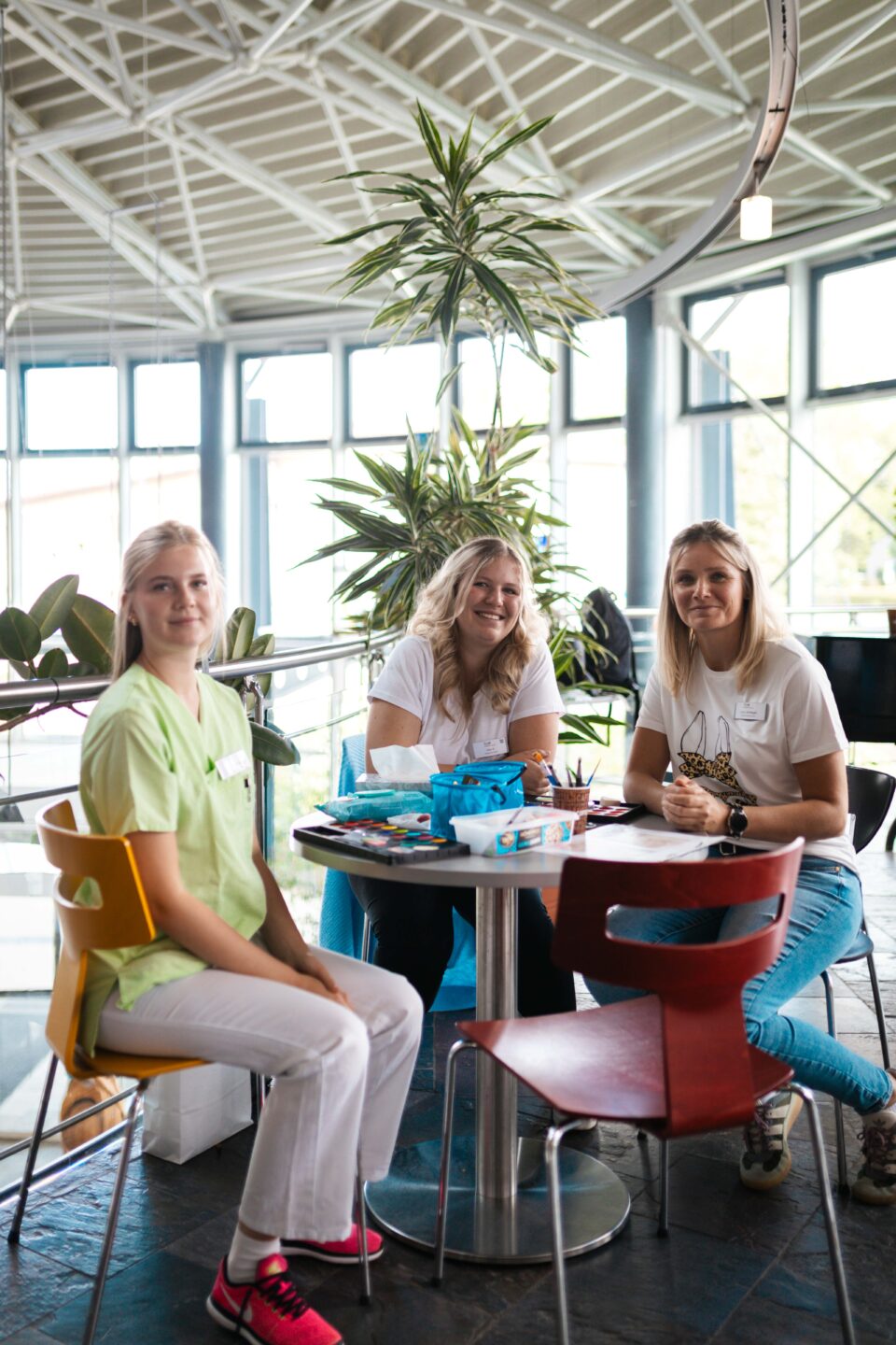 Drei lächelnde Mitarbeiterinnen sitzen an einem runden Tisch mit Kinderschminke und Malutensilien in einem lichtdurchfluteten Foyer.