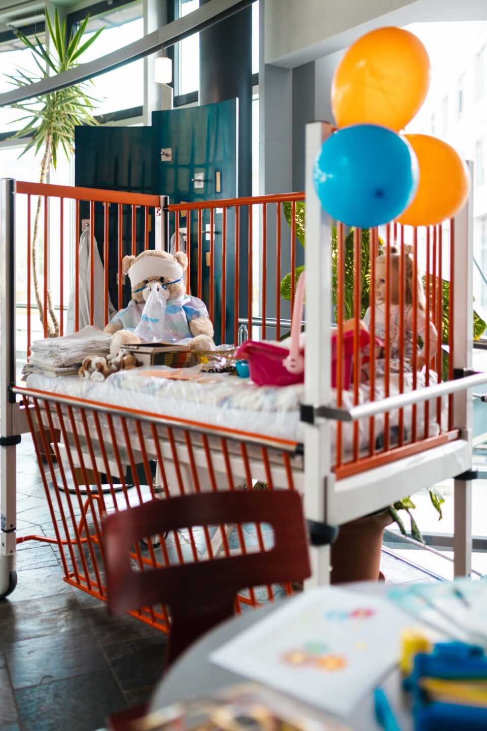 Ein bunt dekoriertes Kinderkrankenhausbett mit einem Teddybären und einer Sauerstoffmaske sowie Luftballons bei einer Veranstaltung.