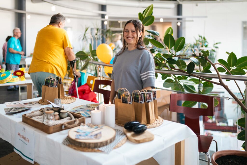 Junge Frau steht lächelnd hinter einem Stand für Physiotherapie und Wellness mit Geschenktüten und Kerzen.