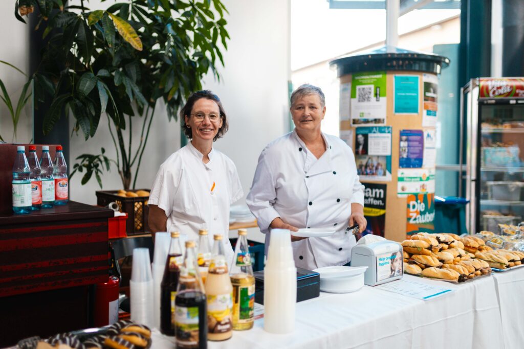 Zwei Mitarbeiterinnen in weißer Arbeitskleidung stehen lächelnd hinter einem Buffet mit belegten Brötchen und Getränken.