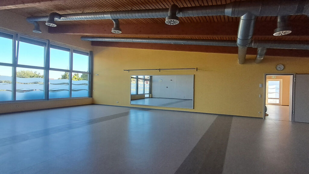 Großzügiger, leerer Kursraum mit gelber Wand, Wandspiegel und moderner Belüftungsanlage an der Holzdecke